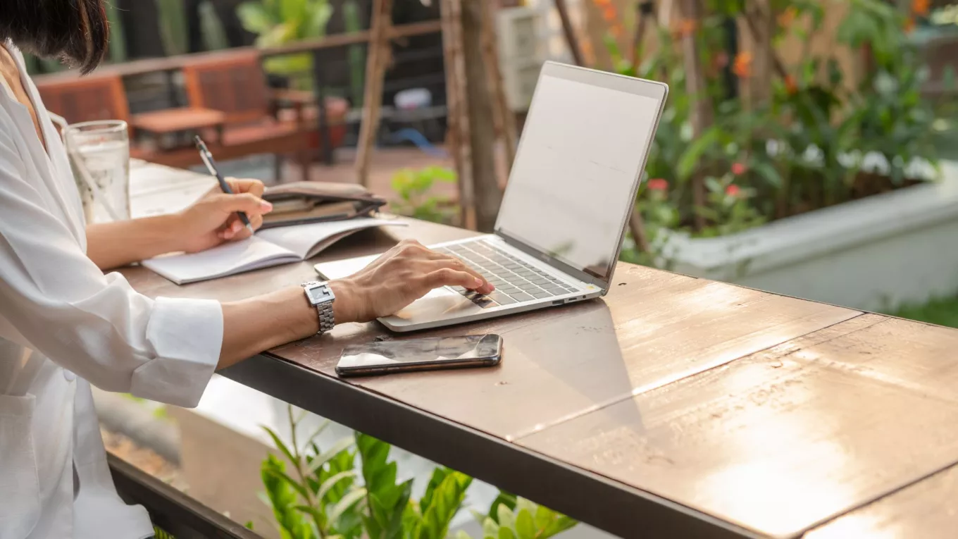 Mujer trabajando de forma remota con una laptop y un cuaderno desde un espacio al aire libre.