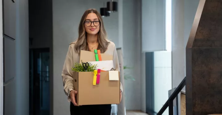 Mujer joven saliendo de su oficina con una caja de pertenencias tras presentar una renuncia voluntaria.