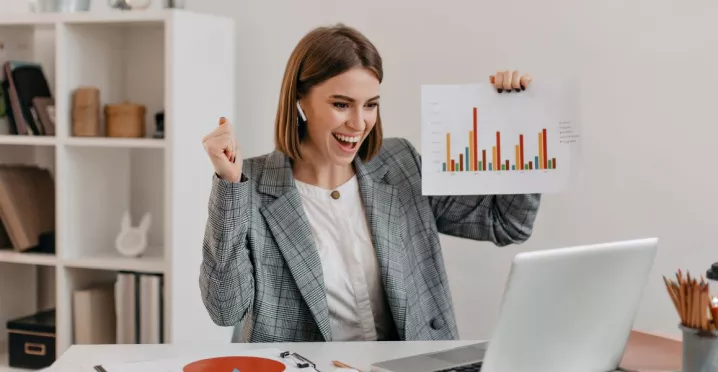 Mujer profesional celebrando resultados frente a su computadora en la oficina, mostrando gráficos de productividad laboral.