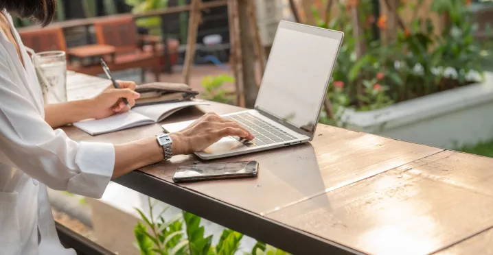 Mujer trabajando de forma remota con una laptop y un cuaderno desde un espacio al aire libre.