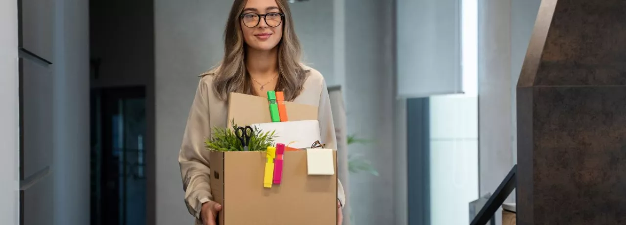 Mujer joven saliendo de su oficina con una caja de pertenencias tras presentar una renuncia voluntaria.