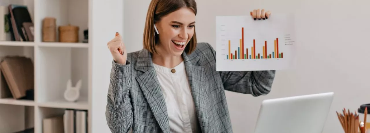 Mujer profesional celebrando resultados frente a su computadora en la oficina, mostrando gráficos de productividad laboral.