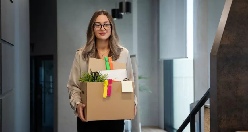 Mujer joven saliendo de su oficina con una caja de pertenencias tras presentar una renuncia voluntaria.