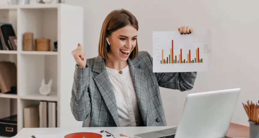 Mujer profesional celebrando resultados frente a su computadora en la oficina, mostrando gráficos de productividad laboral.