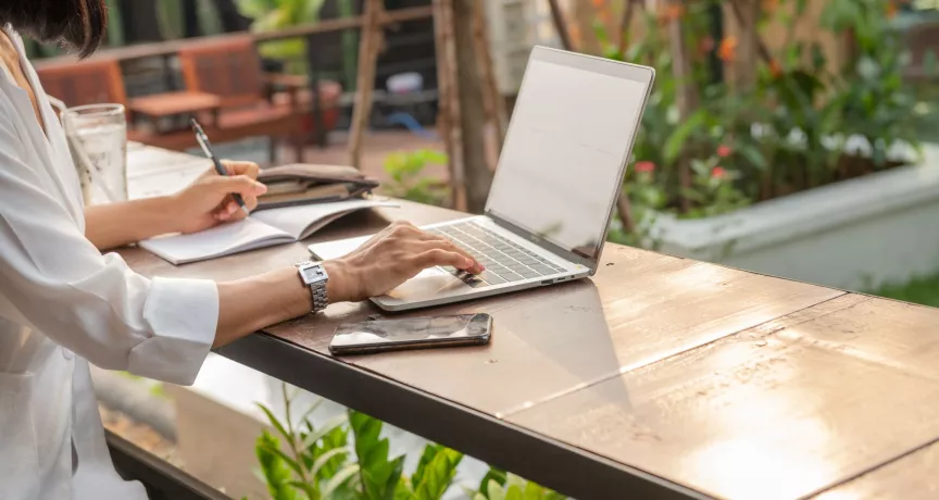 Mujer trabajando de forma remota con una laptop y un cuaderno desde un espacio al aire libre.