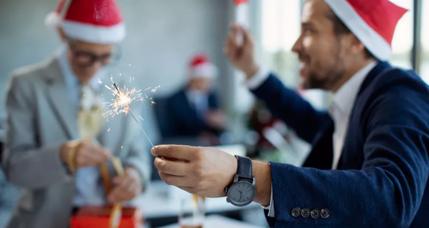 Colaboradores con gorros navideños celebran en la oficina con una bengala encendida y regalos sobre la mesa.