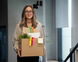 Mujer joven saliendo de su oficina con una caja de pertenencias tras presentar una renuncia voluntaria.
