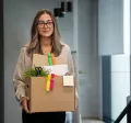 Mujer joven saliendo de su oficina con una caja de pertenencias tras presentar una renuncia voluntaria.
