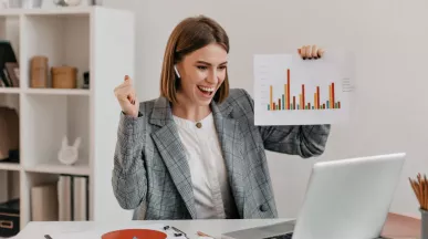 Mujer profesional celebrando resultados frente a su computadora en la oficina, mostrando gráficos de productividad laboral.