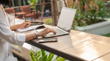 Mujer trabajando de forma remota con una laptop y un cuaderno desde un espacio al aire libre.