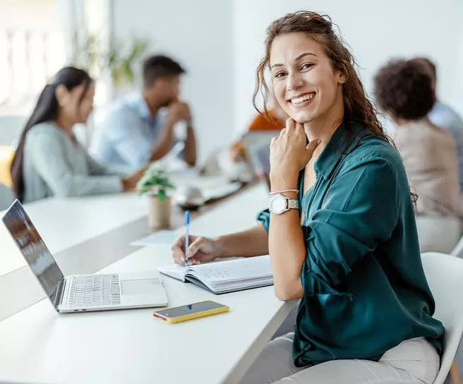 Colaboradora utilizando portátil y tomando notas en un espacio de trabajo moderno, vinculada a programas de Pluxee y Santander.